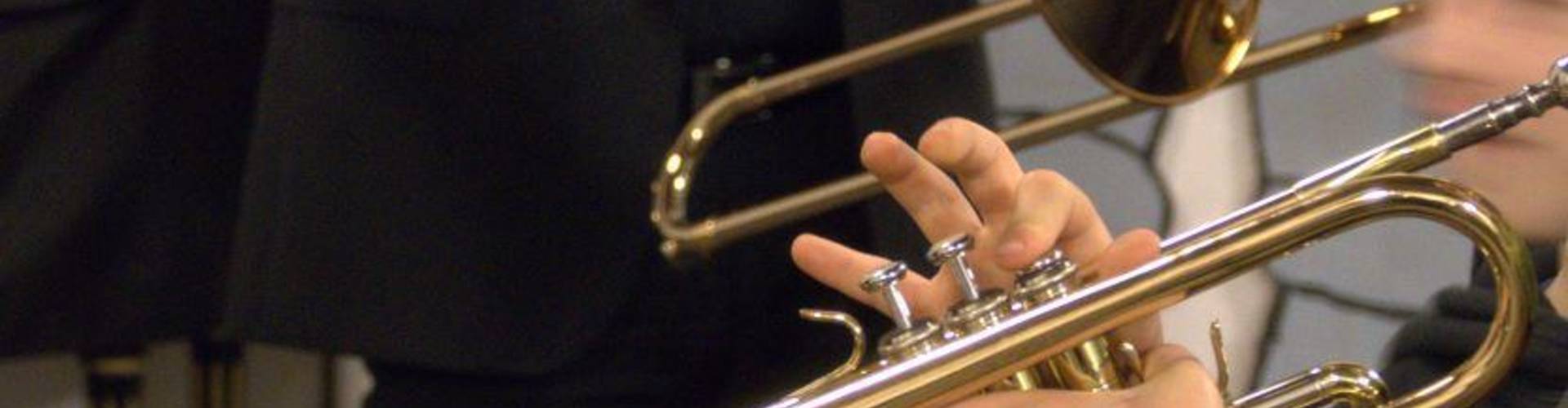 Im Vordergrund sieht man eine Trompete und eine Hand, die das Instrument spielt. Im Hintergrund ist eine Person in schwarzer Kleidung bis zum Oberkörper zu sehen.