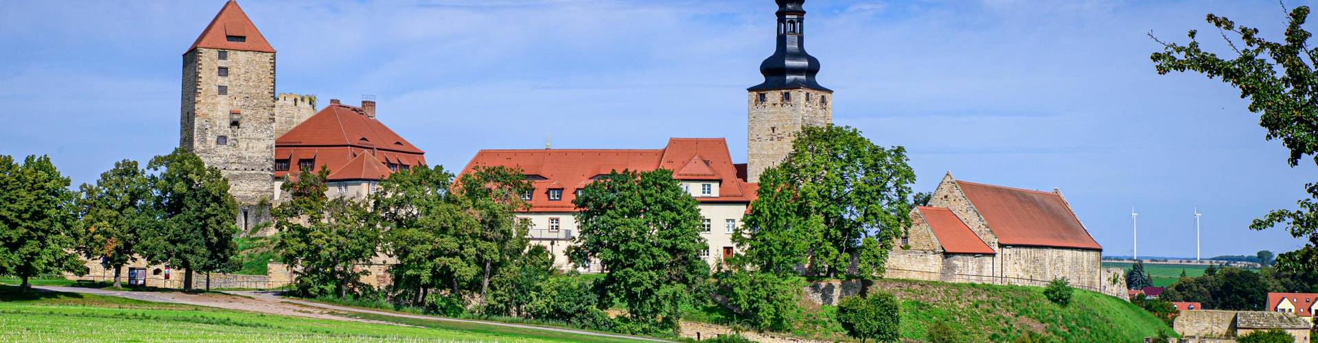 Ein grünes Feld, blauer Himmel, dazwischen Burgturm, Kirchturm und Mauer der Burg Querfurt.