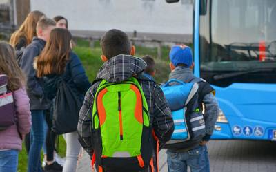 Rechts im Bild ein blauer Bus, davor Mädchen und Jungen mit Ranzen. Sie sind von hinten zu sehen.