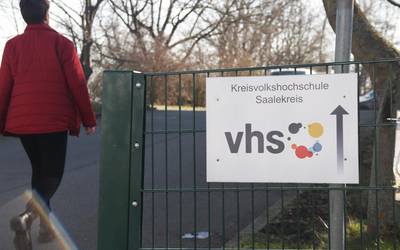 An einem Zaun hängt ein Schild, auf dem in blau Kreisvolkshochschule steht. Links geht eine Frau in einer roten Jacke vorbei.