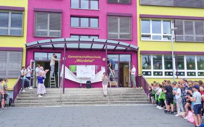 Ein Schulghebäude in rosa und gelb, auf der Treppe ins Gebäude stehen Menschen. Es wird ein Schild freigehängt auf dem steht: Gemeinschaftsschule „Würdetal“. Davor ist der Schulhof frei, links und rechts stehen Schülerinnen und Schüler